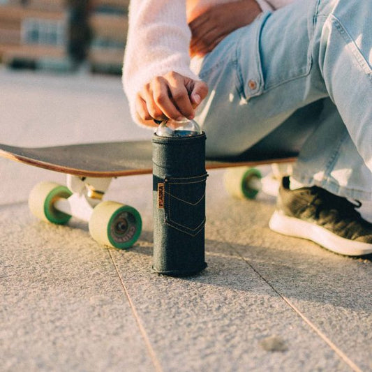 Mano abriendo una botella de vidrio Flaska con funda vaquera, apoyada en el suelo junto a un monopatín.