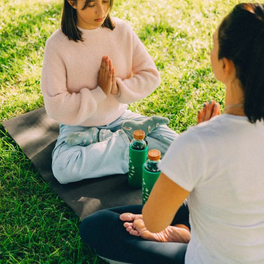 Mujer y niña meditando sobre una esterilla para su bienestar, con dos botellas de vidrio con funda verde en el césped.