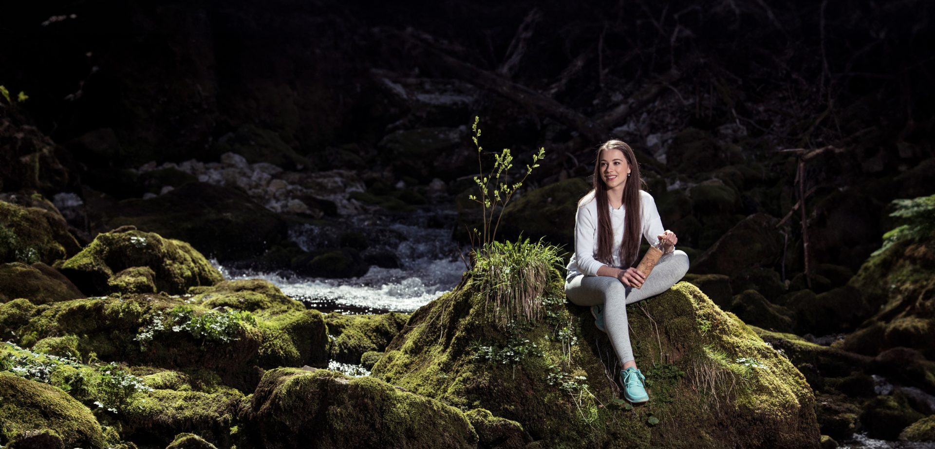 Mujer sonriente sobre una roca con musgo junto a un arroyo, sosteniendo una botella de vidrio para su bienestar.