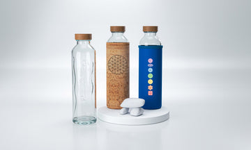 Botellas de agua de cristal Flaska con tapón de madera sobre plataforma circular. Una tiene funda premium de tela de corcho natural y otra funda de neopreno azul.
