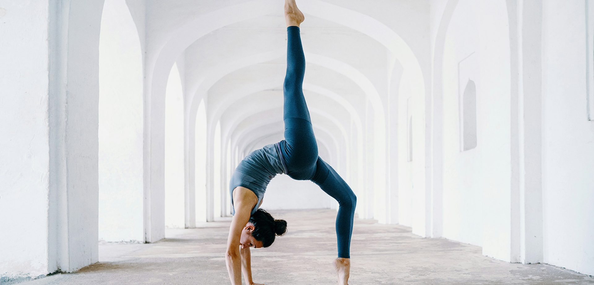 Una persona vestida con ropa deportiva hace una pose de yoga, arqueándose hacia atrás con una pierna levantada, en un pasillo arqueado de color blanco.