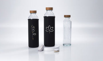 Tres botellas de agua de cristal Flaska: dos con fundas negras de neopreno con diseños de flor de loto y pose de yoga de equilibrio. Todas tienen tapas de madera.