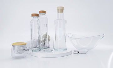 Recipientes de cristal transparente Flaska con tapas de madera y diseños grabados, dispuestos sobre una superficie blanca con formas geométricas y un frutero de cristal.