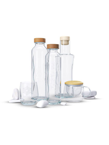 Un grupo de botellas de cristal transparente Flaska con tapas de madera y dos vasos Emoto Flaska, rodeados de piedras blancas, aislados sobre un fondo blanco.